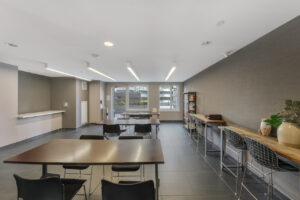 Interior Event space, 4 person tables, bar tables along the perimeter, exit to outdoor rooftop area, gray walls, black tile floor, modern decorations.