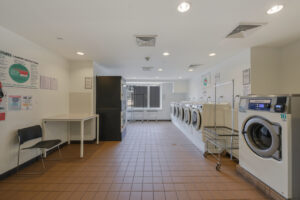 Interior Laundry facilities, tile floor, white walls, rows of washers and dryers, window to the outside, table for folding, multiple laundry carts, seating areas.