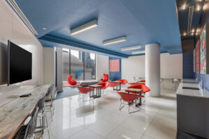 interior Lounge area, large TV, long table in front of tv with 4 seats, long countertop with sink, lounge chairs with ottomans in front of large floor to ceiling windows, red chairs across eachother with small tables through the room, white tile floor, contemporary art on the walls, stunning view.