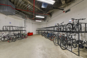 Interior bike storage room, multiple large bike racks, exposed industrial ceiling, white walls, racks sectioned by letters, red storage chest, concrete floor.