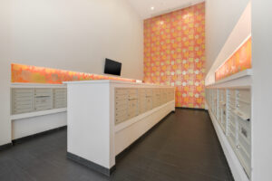 Interior Mail Room, pink yellow and orange wallpaper the rest white walls, 4 rows of mailboxes, black tile floor, TV in room, large ceiling.