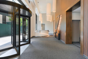 Interior lobby entrance, wood walls near elevator, large 7 dekalb signage upon entering, front desk to the left of entrance, gray carpeting, large overhead light installation, revolving doors.
