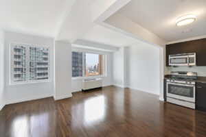 Interior Unit Living Room, wood floors, open floor plan, stainless steel appliances in kitchen, ac unit near window, 21 windows with views of downtown, white walls.